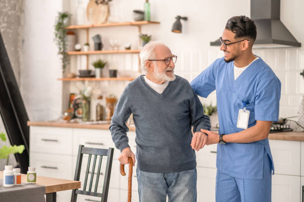 Smiling friendly in-home male nurse in uniform supporting an old man with a walking stick